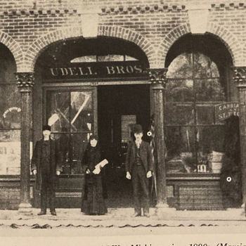 Three people stand outside a brick storefront with arched windows labeled "Udell Bros." in an old black-and-white photo, conveying a historical atmosphere.