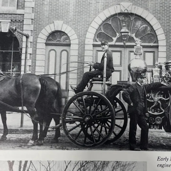 Vintage photo of a horse-drawn fire engine with two firefighters in uniform. One sits on the wagon, while the other stands beside it. Brick building in background.