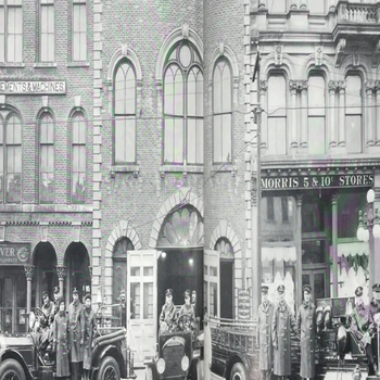 Historic black-and-white photo of firefighters in long coats standing with fire trucks outside a brick fire station, conveying a vintage urban scene.