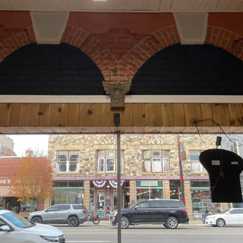 Street view through ornate arches with rustic wood and brick design, showing cars parked in front of a stone building adorned with American flags.