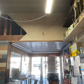 A hardware store interior with high shelves filled with paint cans on the right. A large, glowing overhead light illuminates a rustic wooden counter below.