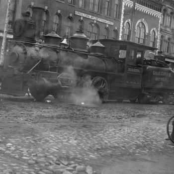 Historic black and white photo of a steam locomotive on cobblestone street, with smoke billowing, in front of ornate brick buildings. Vintage urban scene.