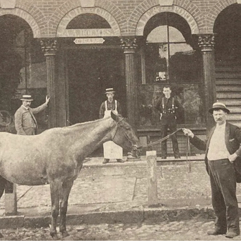 A vintage photo shows four men in hats and suits, posing with a horse on a cobblestone street. They're in front of a brick building with arched doorways.