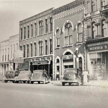 A black-and-white photo of a historic street with vintage cars parked in front of ornate brick buildings. Signs display "Arcade" and "Morris 5." A few people are walking nearby.