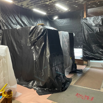 Room with black and white plastic coverings draping over furniture, creating a makeshift barrier. Overhead lights highlight a renovation or construction scene.