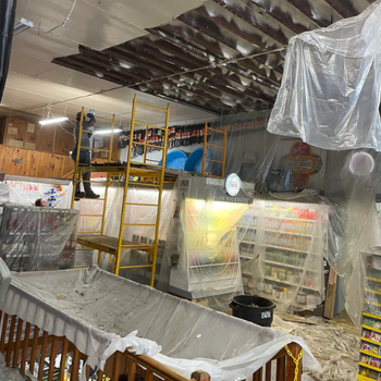 A store ceiling collapses with debris scattered below. Scaffolding and plastic sheets cover shelves. A worker in safety gear stands on the scaffolding.