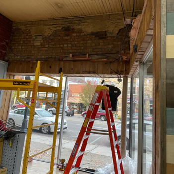 A store interior under renovation with exposed brick and wooden beams. A red ladder and tools are in the foreground, and a street view is visible outside.