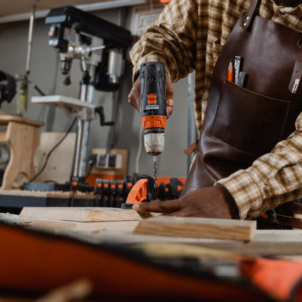 Power ToolsA person wearing a plaid shirt and leather apron uses a power drill on wood in a workshop. Tools and equipment are visible in the background.