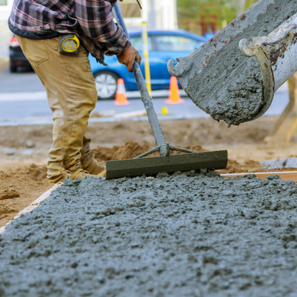 ConcreteA worker in a plaid shirt and beige pants spreads wet concrete with a tool. A concrete mixer pours fresh mix. Traffic cones and a car are in the background.